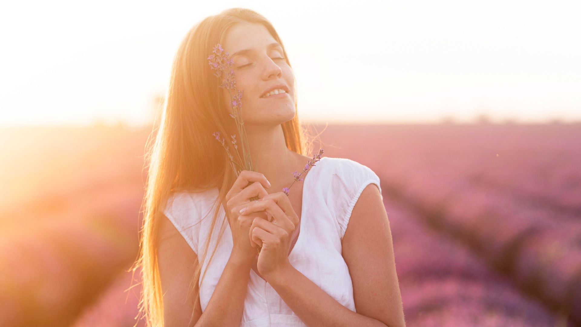 Mulher com uma blusa branca em um jardim com o rosto iluminado pelo sol, olhando para cima em tom de estar fazendo uma oração para acalmar o coração.
