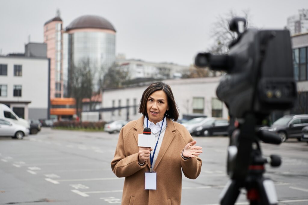 A multiracial journalist in a beige coat stands in a parking lot, confidently delivering a news report.