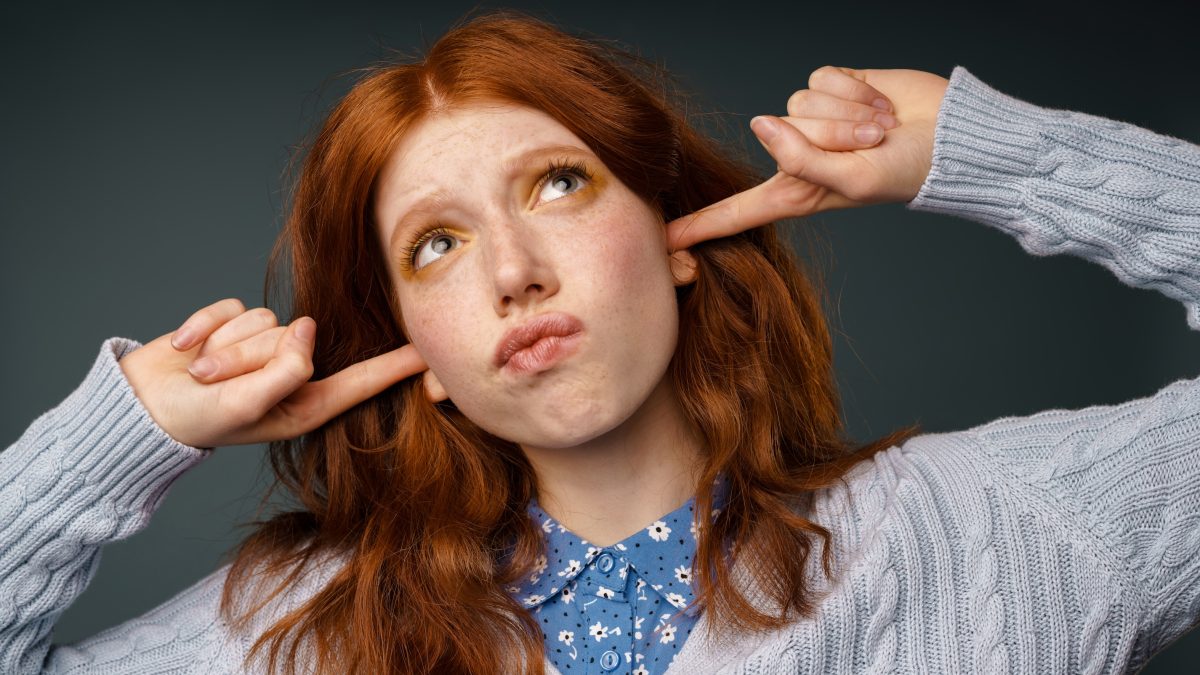 Young ginger displeased woman looking upward while plugging her ears