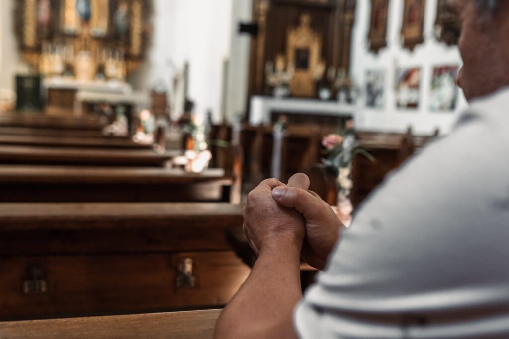 middle-aged man, in casual clothes, sits on bench in Catholic church, alone, prays with eyes closed