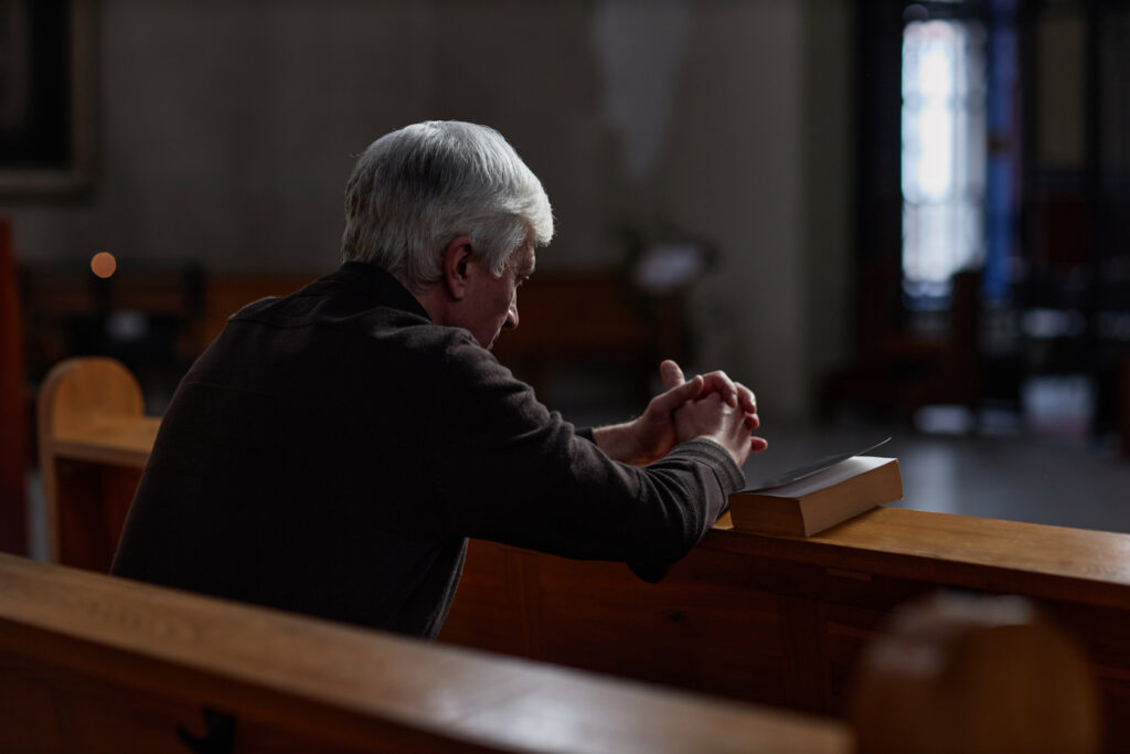 Rear view of senior man sitting on the bench with Bible and praying in old church